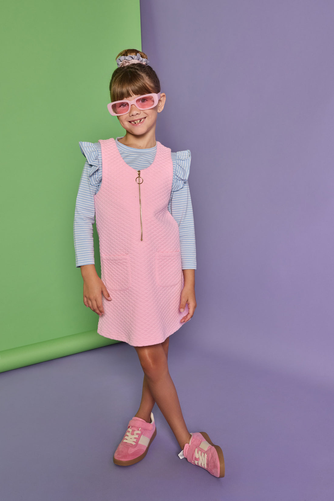 Model can be seen wearing a blue and white stripe long sleeve knit top with ruffles at the sleeves paired under a bubblegum pink quilted jumper. Jumper features quarter zip zipper down front and pockets. 