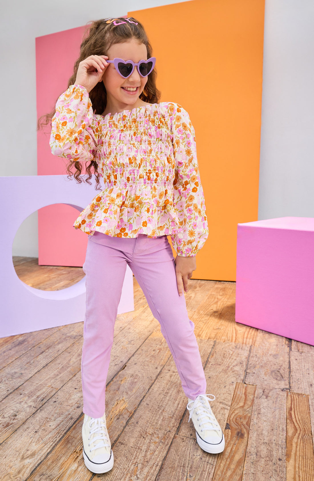 Young girl in a colorful outfit with sunglasses standing in front of colorful geometric shapes.