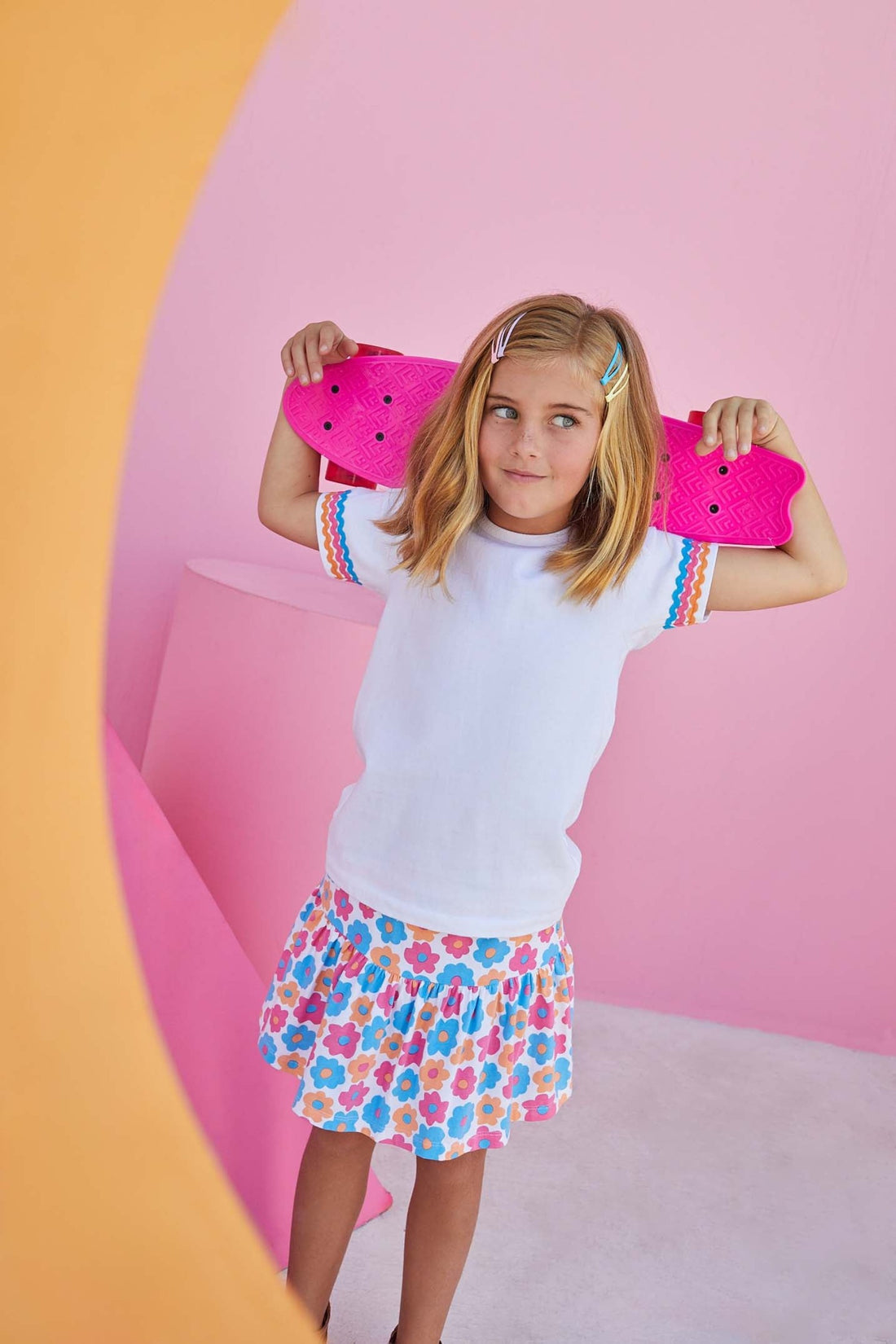Our model is holding a skateboard behind her head and wearing our Ric Rac essential tee which features three layers of ric rac detailing on the short sleeves in bright blue, pink, and orange. 