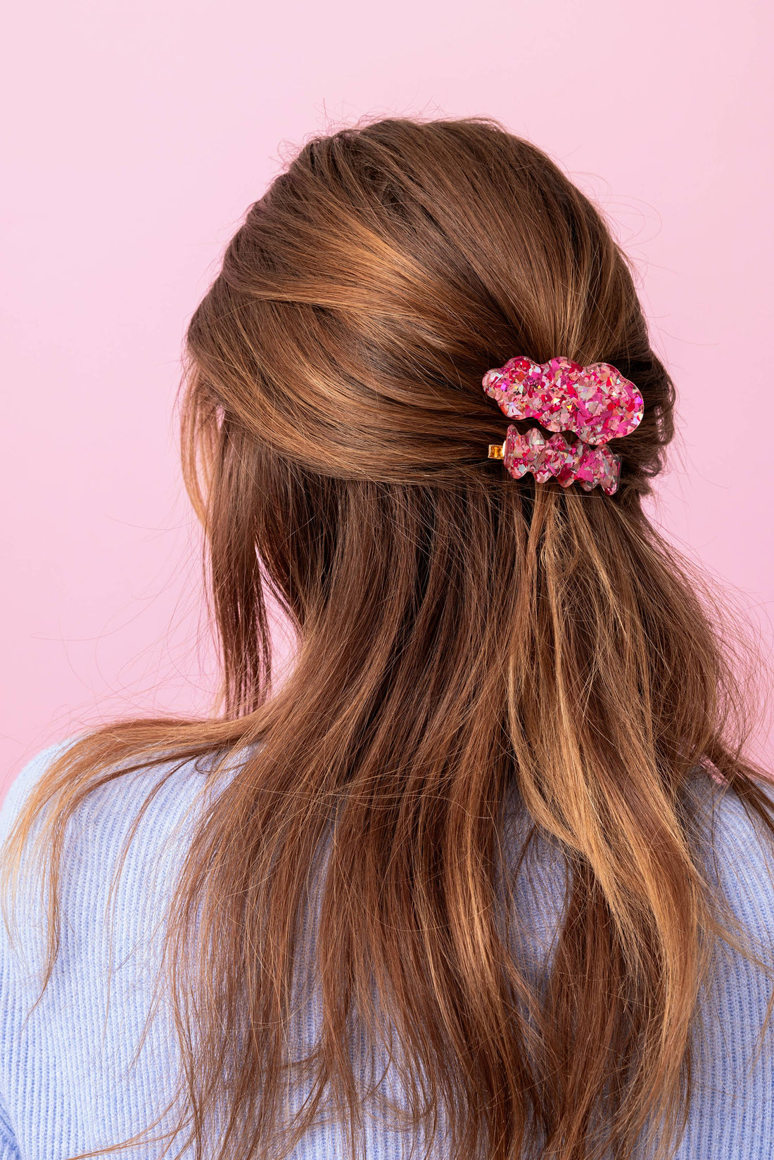 Woman with long brown hair styled in a half-up, half-down style with a pink floral hair accessory against a pink background.