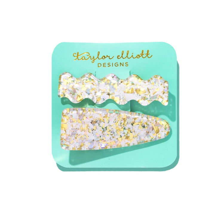 Two decorative hair clips with gold and white glitter on a teal card.